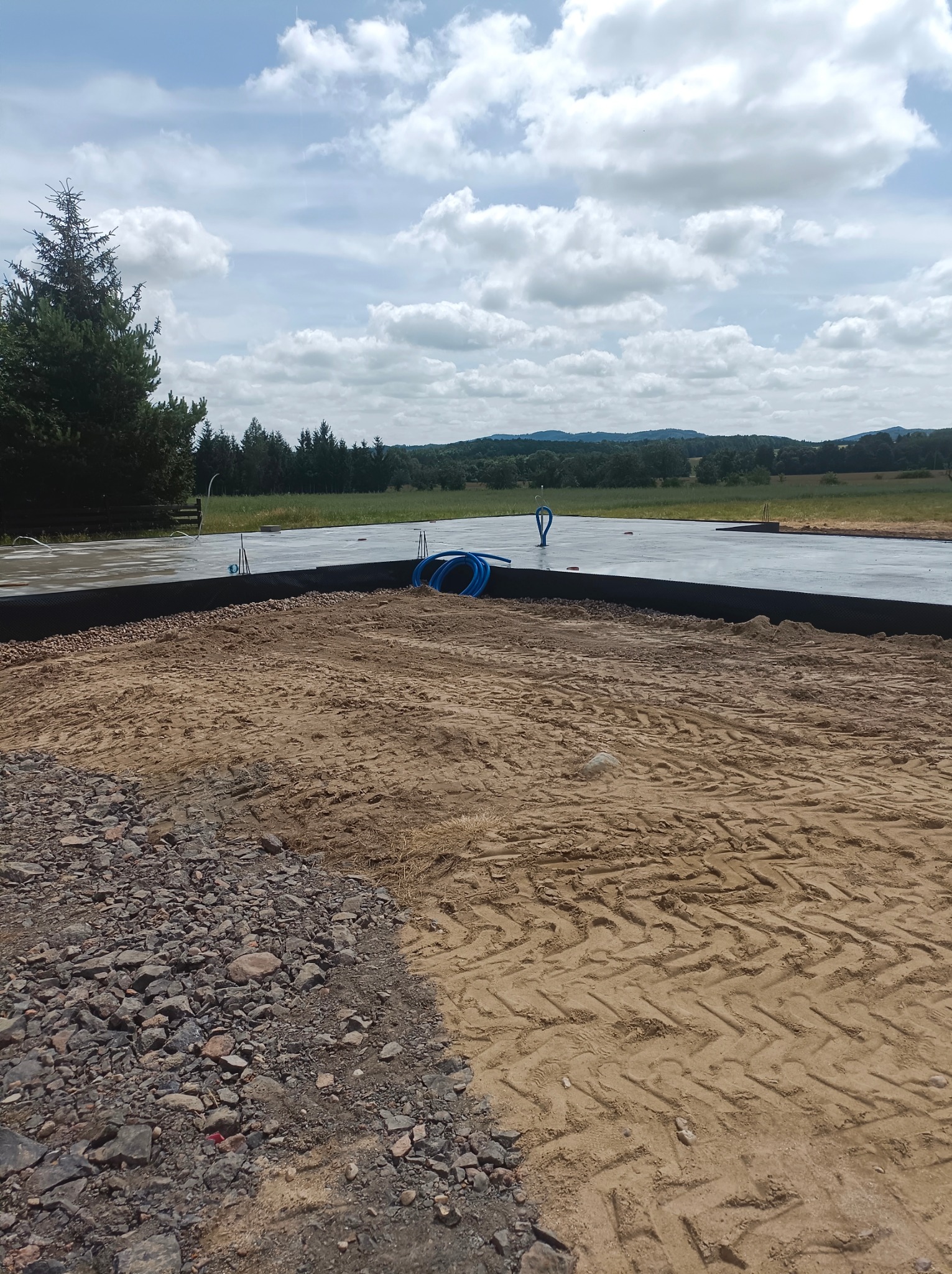 Wylana płyta fundamentowa na przygotowanym terenie w Sędziszowej, widoczne niebieskie rury instalacyjne i czarna folia hydroizolacyjna, teren częściowo zasypany piaskiem z widocznymi śladami opon.