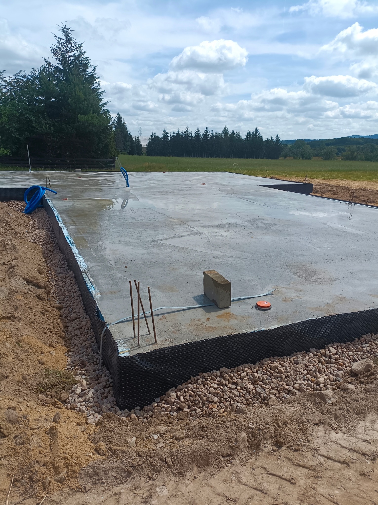 Świeżo wylana, mokra płyta fundamentowa z wystającymi prętami zbrojeniowymi, zabezpieczona folią kubełkową i obsypana kamieniami, na tle krajobrazu z drzewami i pochmurnym niebem.