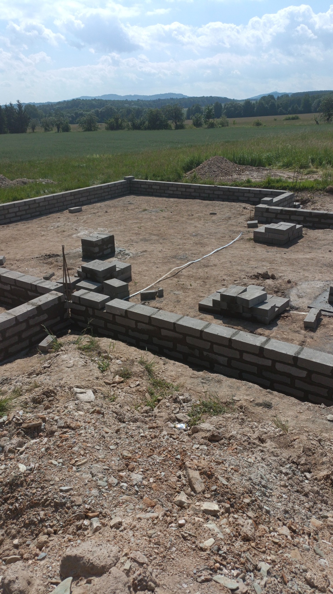 Wykopane fundamenty z częściowo wymurowanymi ścianami z betonowych bloczków, widok na zielone pole i las w oddali.