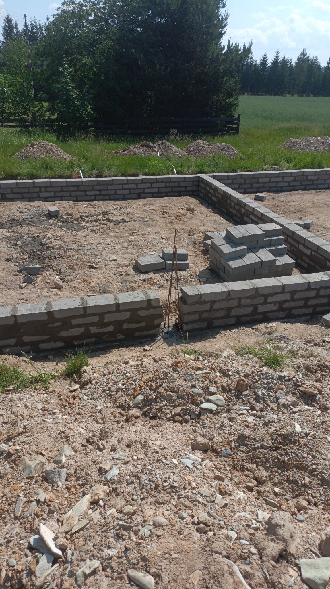 Wykop fundamentowy z częściowo wymurowanymi ścianami z bloczków betonowych, widoczne pręty zbrojeniowe i stos bloczków, w tle zieleń.