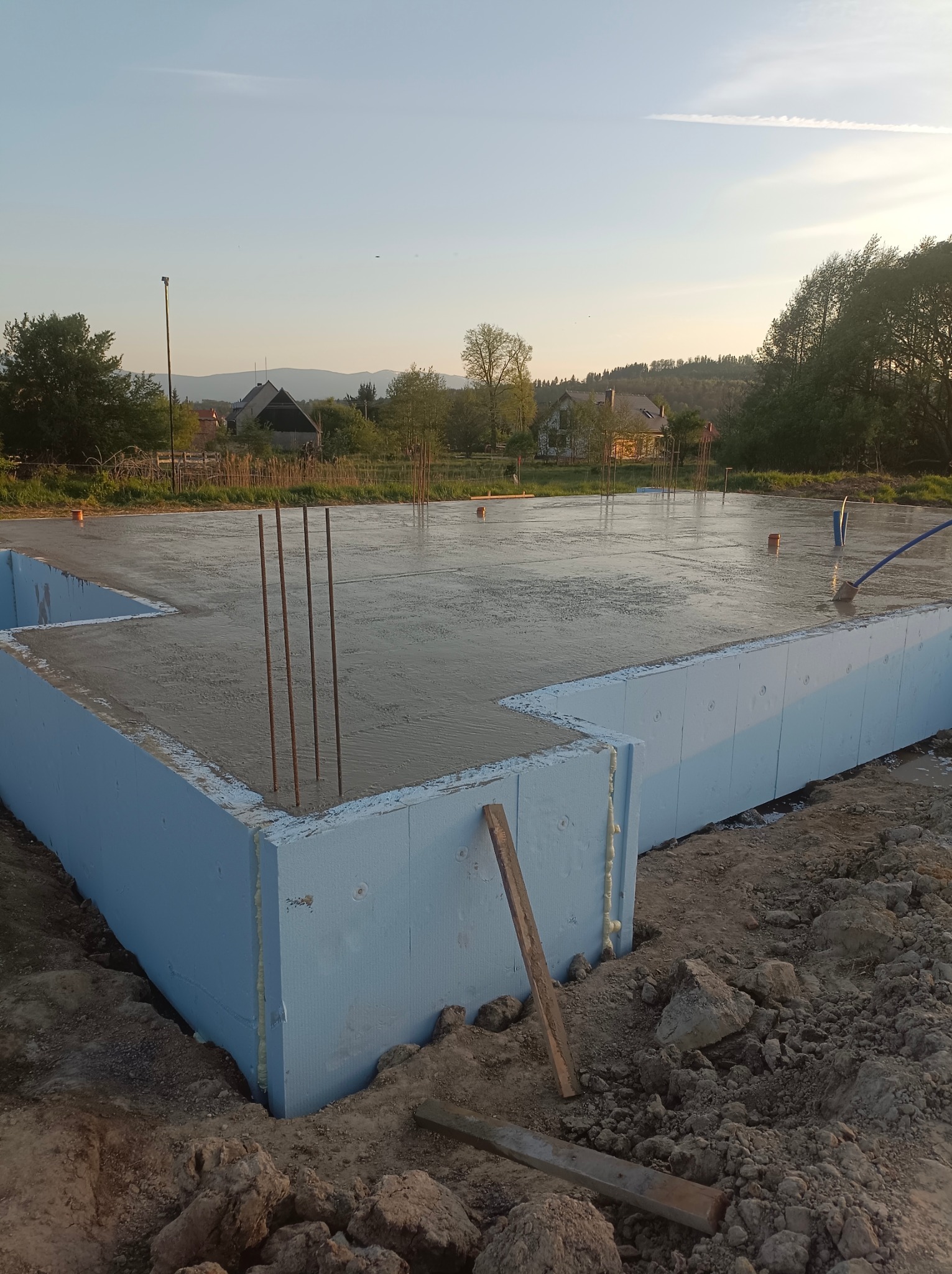 Wylany fundament z wystającymi prętami zbrojeniowymi, zabezpieczony niebieskim styropianem, na tle wiejskiego krajobrazu o zmierzchu.