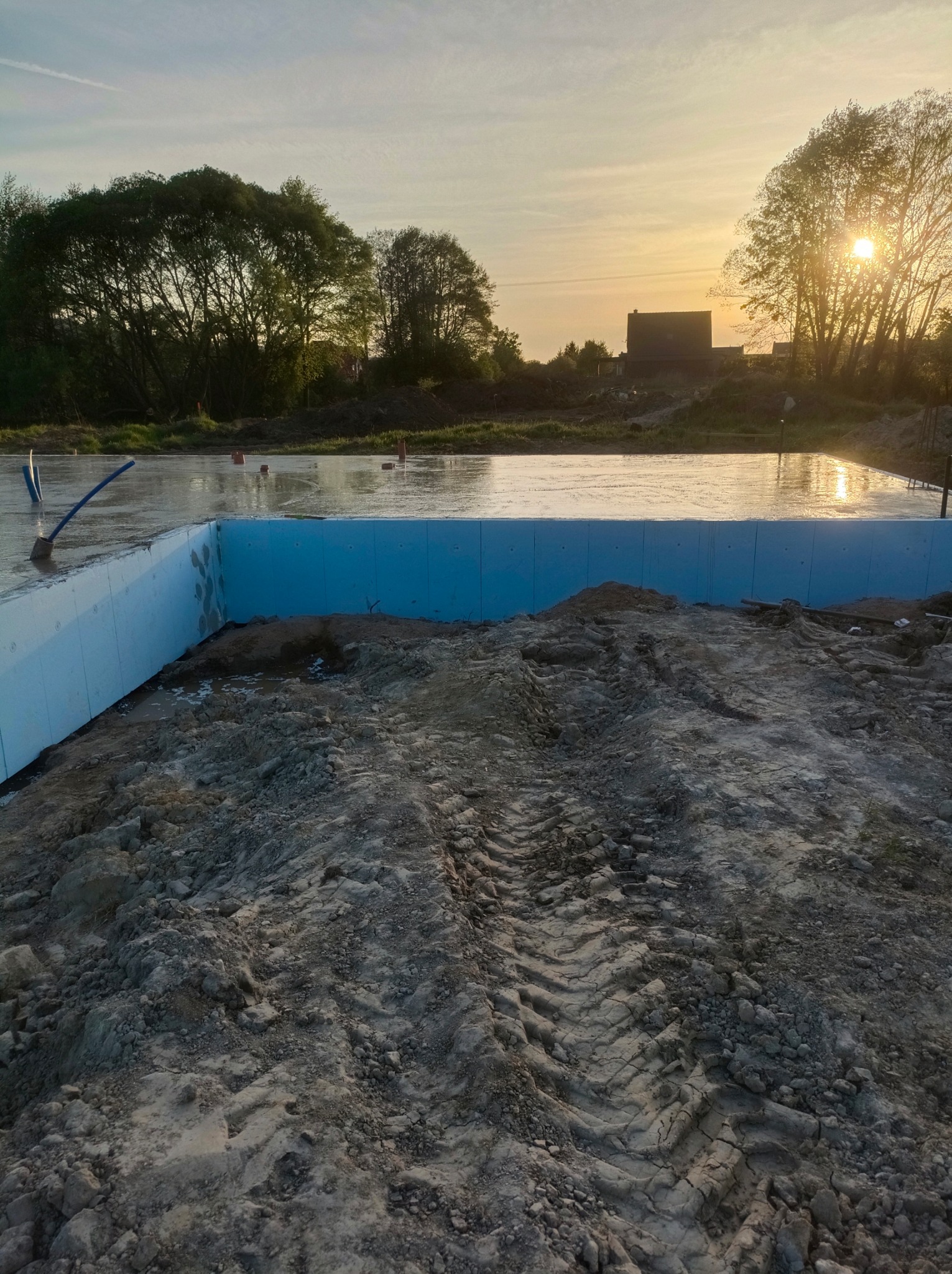 Wylany fundament z izolacją pionową w postaci niebieskich płyt styropianowych, w tle zachodzące słońce i zarys budynku.