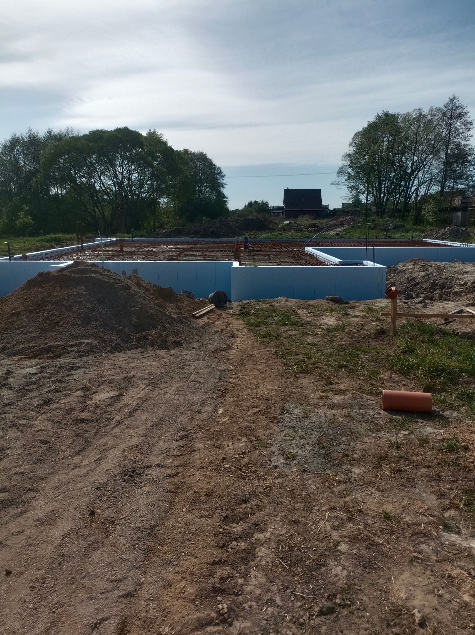 Przygotowanie terenu pod fundamenty domu jednorodzinnego z widocznym zbrojeniem i izolacją termiczną, na pierwszym planie hałda ziemi i pomarańczowa rura.