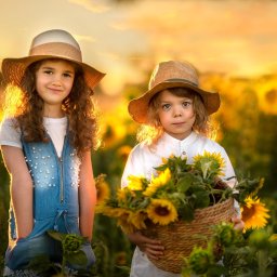 Beata Orcholska Photography - Dwie dziewczynki w kapeluszach stoją na polu słoneczników podczas zachodu słońca, jedna trzyma kosz pełen słoneczników, druga ma ręce w kieszeniach dżinsowego kombinezonu ozdobionego perełkami.