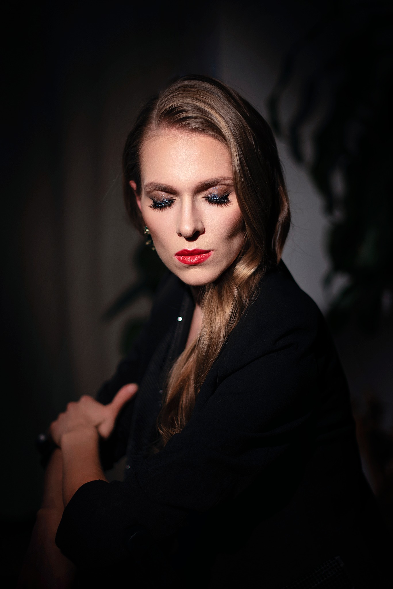 Portret kobiety z błyszczącym makijażem oczu i czerwonymi ustami, ubrana w czarną marynarkę, z zamyślonym spojrzeniem skierowanym w dół, oświetlona bocznym światłem.
