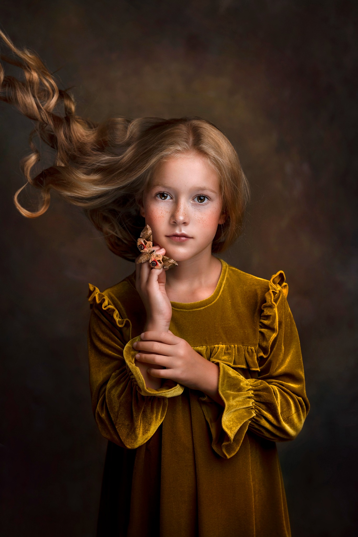 Portret dziewczynki z rozpuszczonymi blond włosami i piegami, trzymającej sztucznego motyla na dłoni, w sukience z żółtego weluru na ciemnym tle.