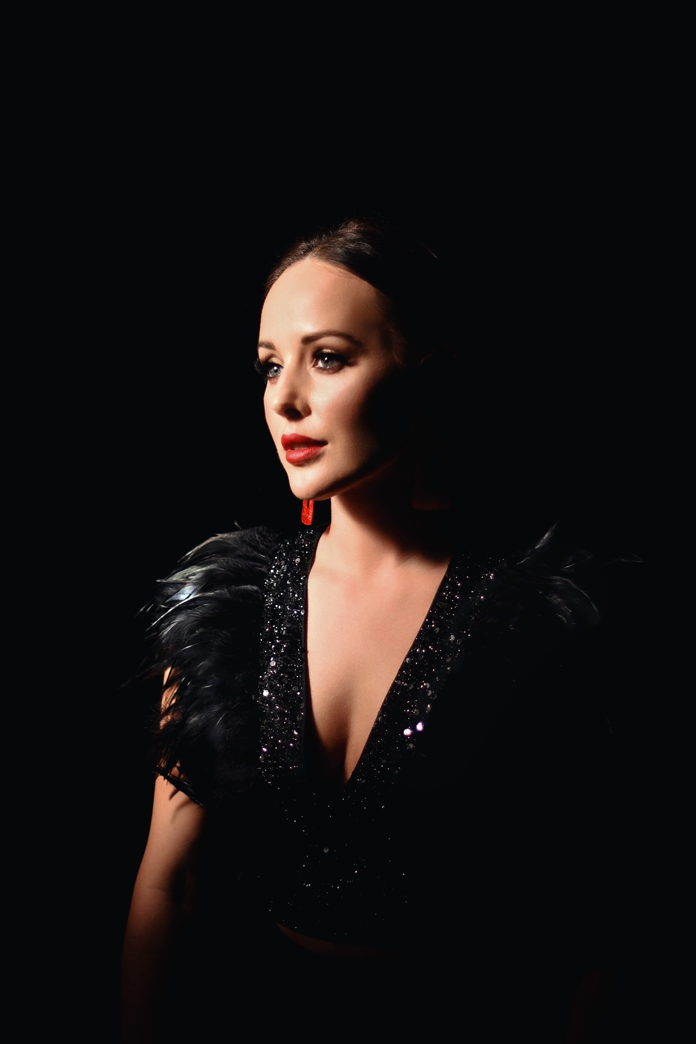 Portret kobiety w czarnej, błyszczącej sukience z piórami na ramionach, czerwone usta i kolczyki, oświetlenie boczne, ciemne tło.