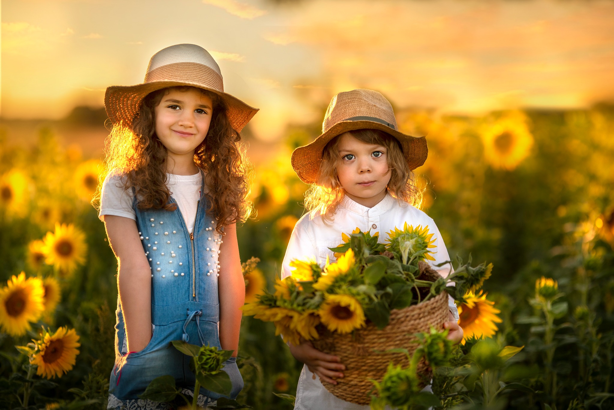 Dwie dziewczynki w kapeluszach stoją na polu słoneczników podczas zachodu słońca, jedna trzyma kosz pełen słoneczników, druga ma ręce w kieszeniach dżinsowego kombinezonu ozdobionego perełkami.