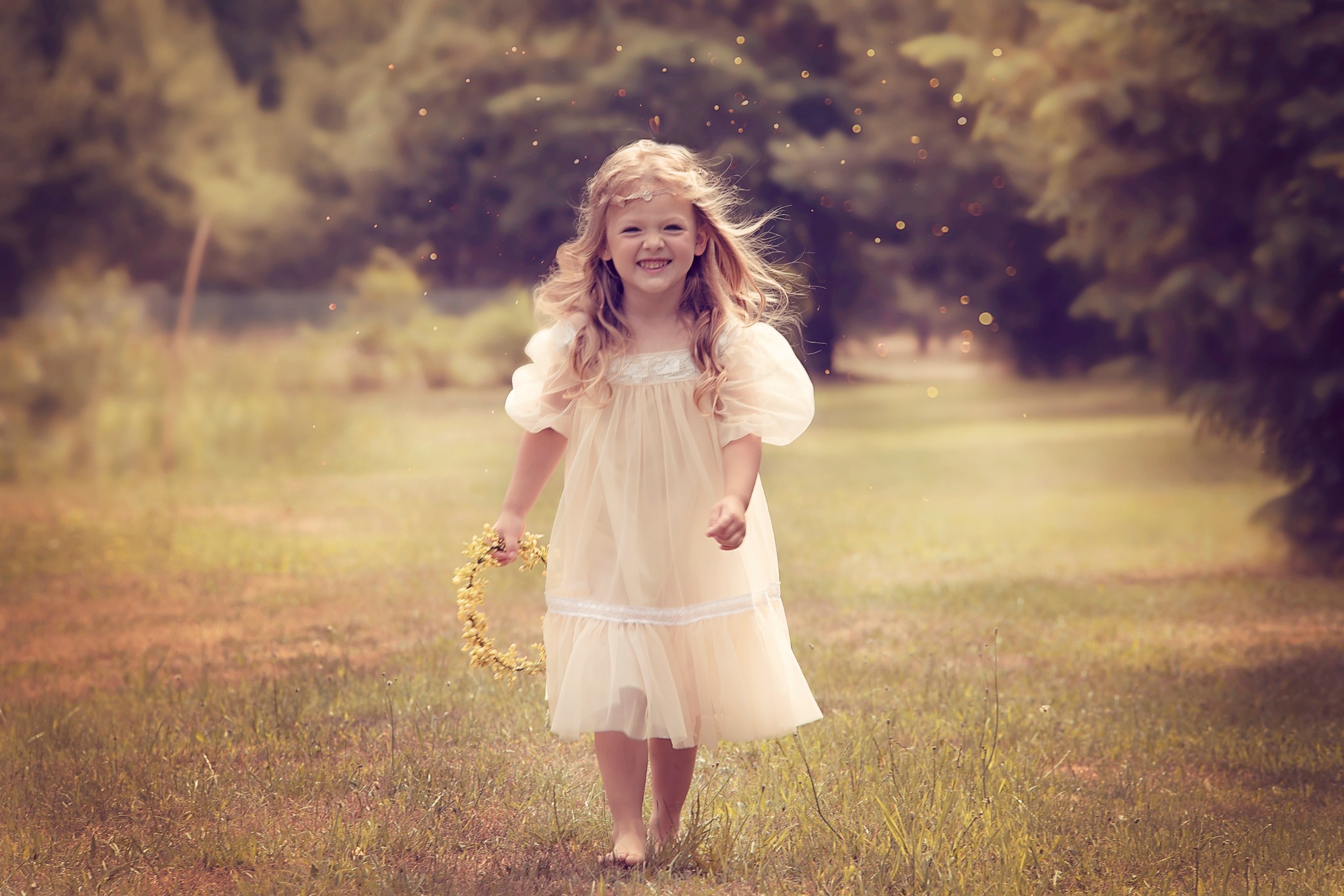 Uśmiechnięta dziewczynka z wiankiem biegnie boso po trawie w zwiewnej sukience, złote refleksy światła w tle drzew