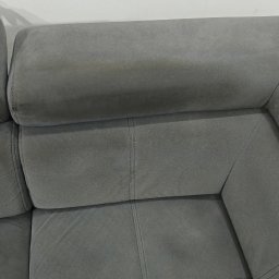 Maxim Marcin Puzyniak - Fragment szarej, tapicerowanej kanapy z widocznymi szwami i zagłówkiem na tle białej ściany.