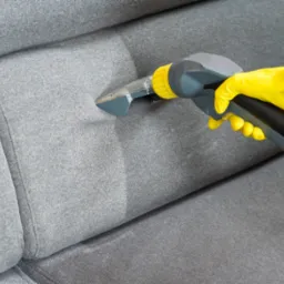 Czyszczenie fragmentu szarej sofy przy użyciu odkurzacza piorącego, widoczna różnica w odcieniu tkaniny po czyszczeniu, osoba w żółtych rękawicach obsługuje urządzenie.