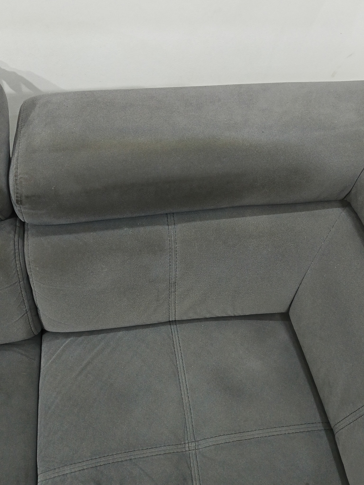 Fragment szarej, tapicerowanej kanapy z widocznymi szwami i zagłówkiem na tle białej ściany.