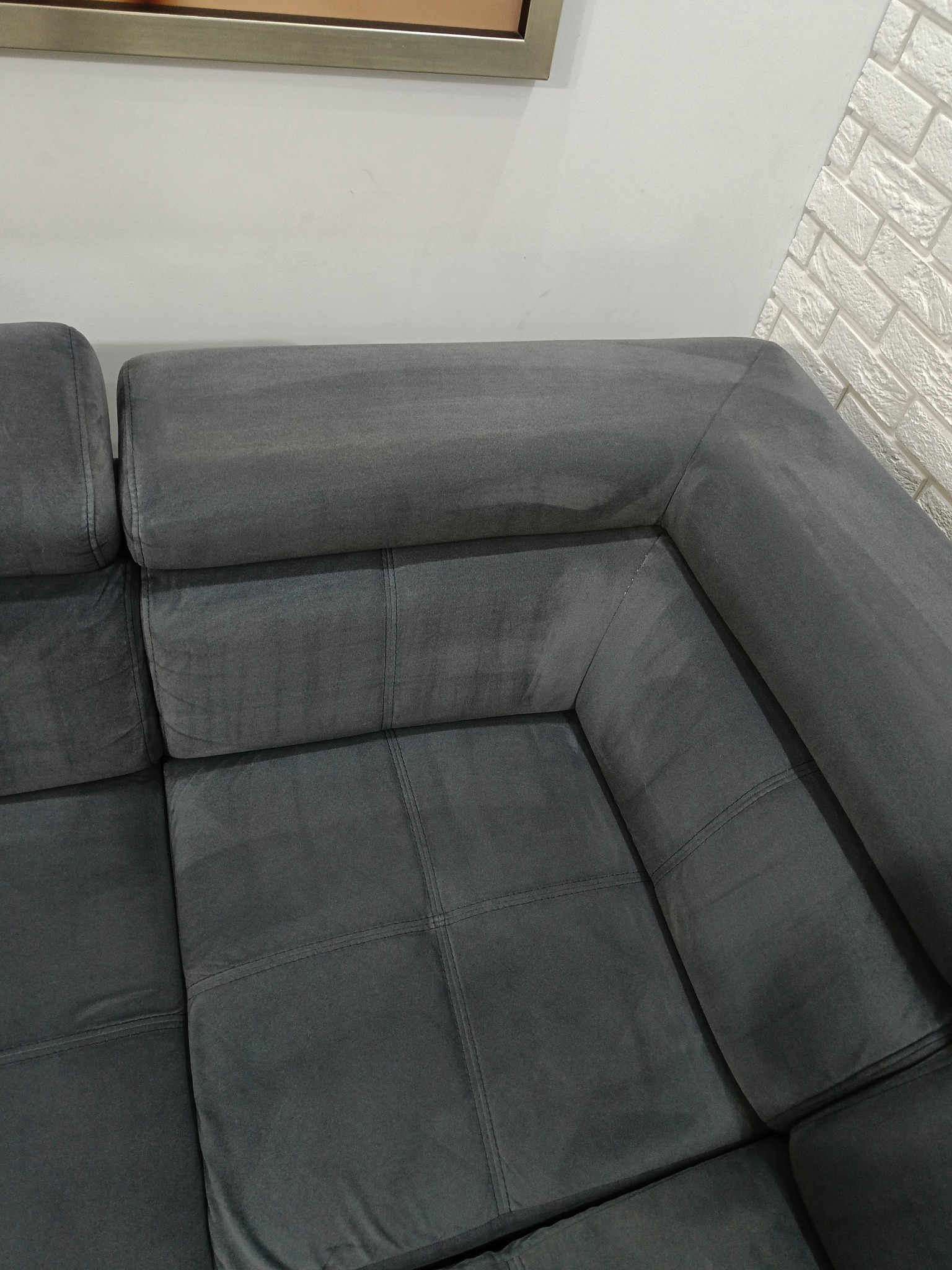 Fragment szarej, narożnej kanapy z widocznymi śladami po czyszczeniu, na tle białej ściany i dekoracyjnej cegły.