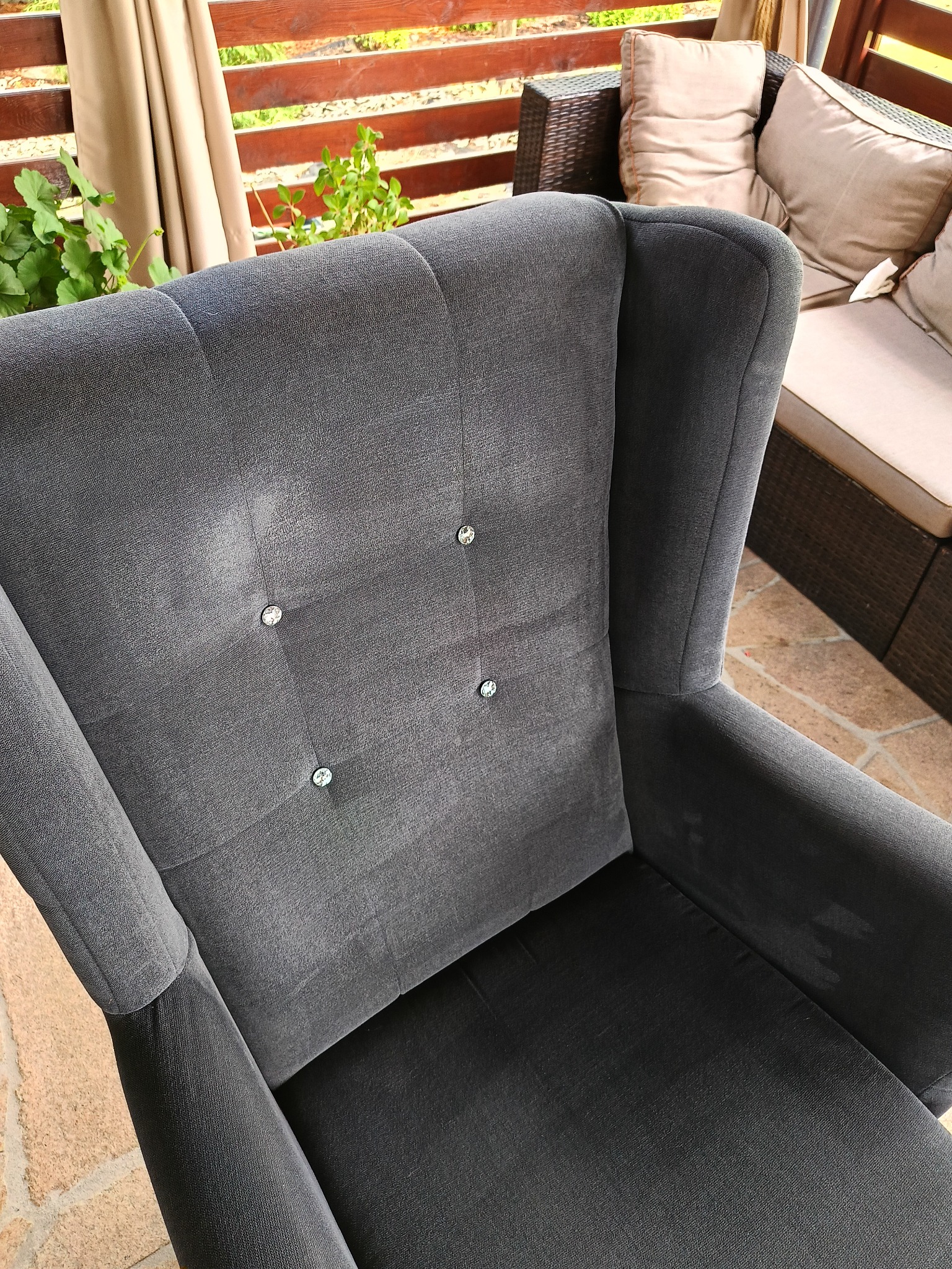 Szary fotel typu uszak z welurową tapicerką i dekoracyjnymi guzikami, widoczne plamy na oparciu, w tle meble ogrodowe i drewniana balustrada.