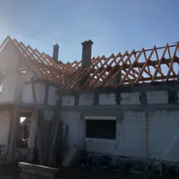 Surowy budynek w trakcie budowy z widoczną drewnianą konstrukcją dachu, dwa kominy murowane, jasne niebo w tle i promienie słoneczne.