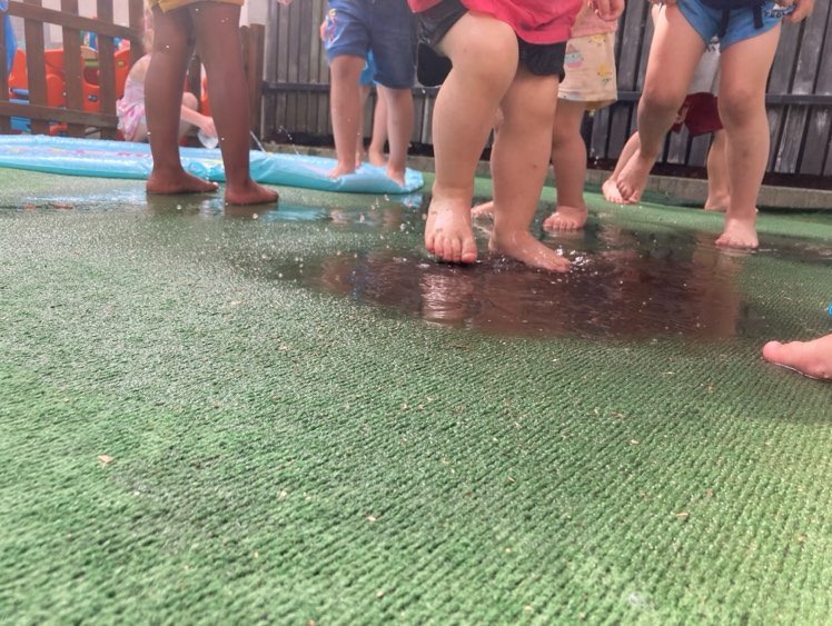 Dzieci bawią się w wodzie na zielonej sztucznej trawie, widoczne stopy i nogi w różnym wieku, w tle basenik.