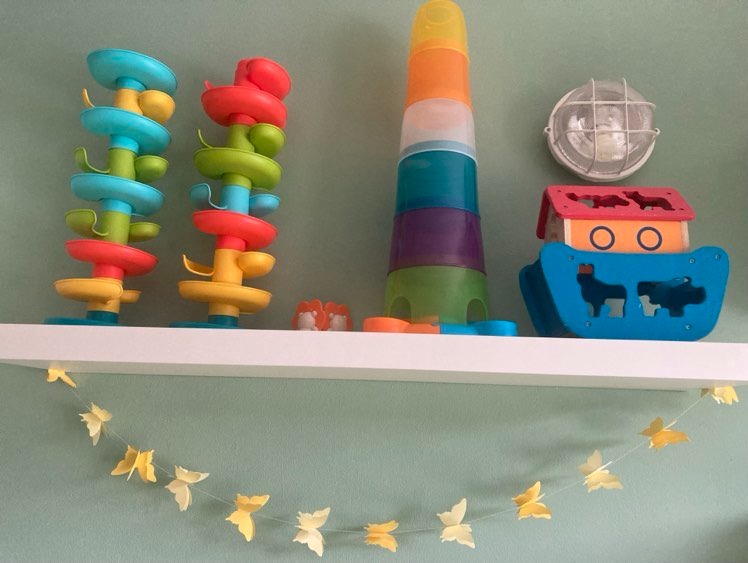Kolorowe zabawki edukacyjne i drewniana arka Noego na białej półce, ozdobione girlandą z papierowych motyli na tle zielonej ściany.