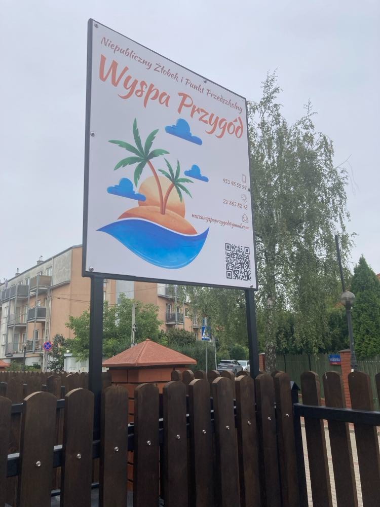 Szyld Niepublicznego Żłobka i Punktu Przedszkolnego 'Wyspa Przygód' z grafiką wyspy z palmami i chmurkami, umieszczony na metalowym stelażu przed budynkiem i drewnianym płotem.