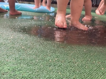 Dziecięce stopy brodzące w płytkiej wodzie na zielonej, teksturowanej powierzchni, prawdopodobnie sztucznej trawie, z fragmentem niebieskiego dmuchanego baseniku w tle.