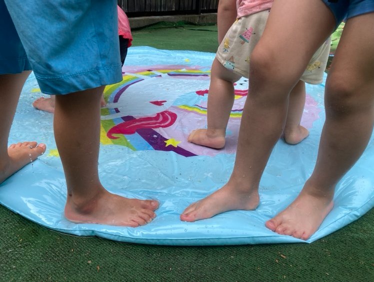 Dzieci bawią się na dmuchanej, wodnej macie z motywem jednorożca, widoczne stopy i nogi.