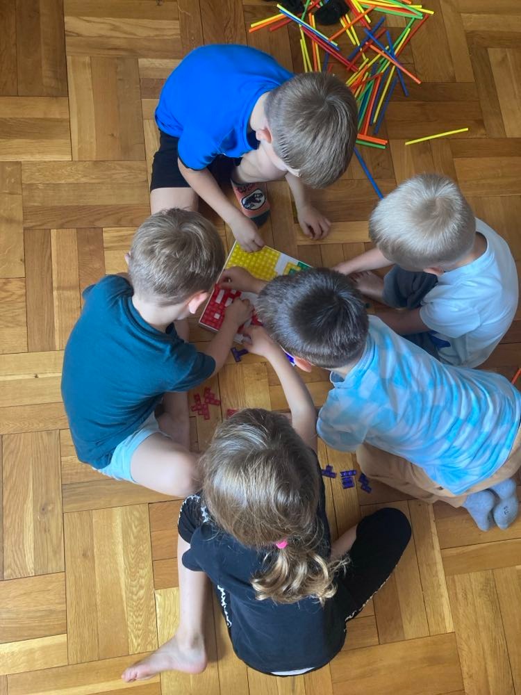 Pięcioro dzieci siedzi w kręgu na parkiecie i wspólnie gra w grę planszową z kolorowymi elementami, wokół leżą patyczki do liczenia.