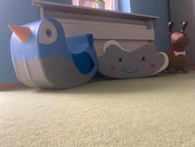 Pokój dziecięcy z miękkimi zabawkami do siedzenia w kształcie niebieskiego ptaka, szarej chmurki z uśmiechem i brązowego pieska z kością na szyi, ustawionymi na zielonym dywanie.