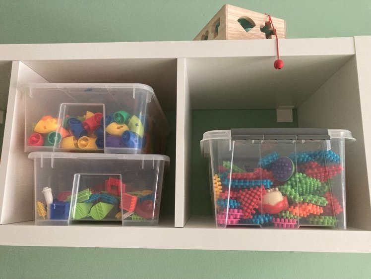 Półka z zabawkami edukacyjnymi w przezroczystych pojemnikach, w tle zielona ściana, na górnej półce drewniana kostka edukacyjna z czerwonym sznurkiem i kulką.