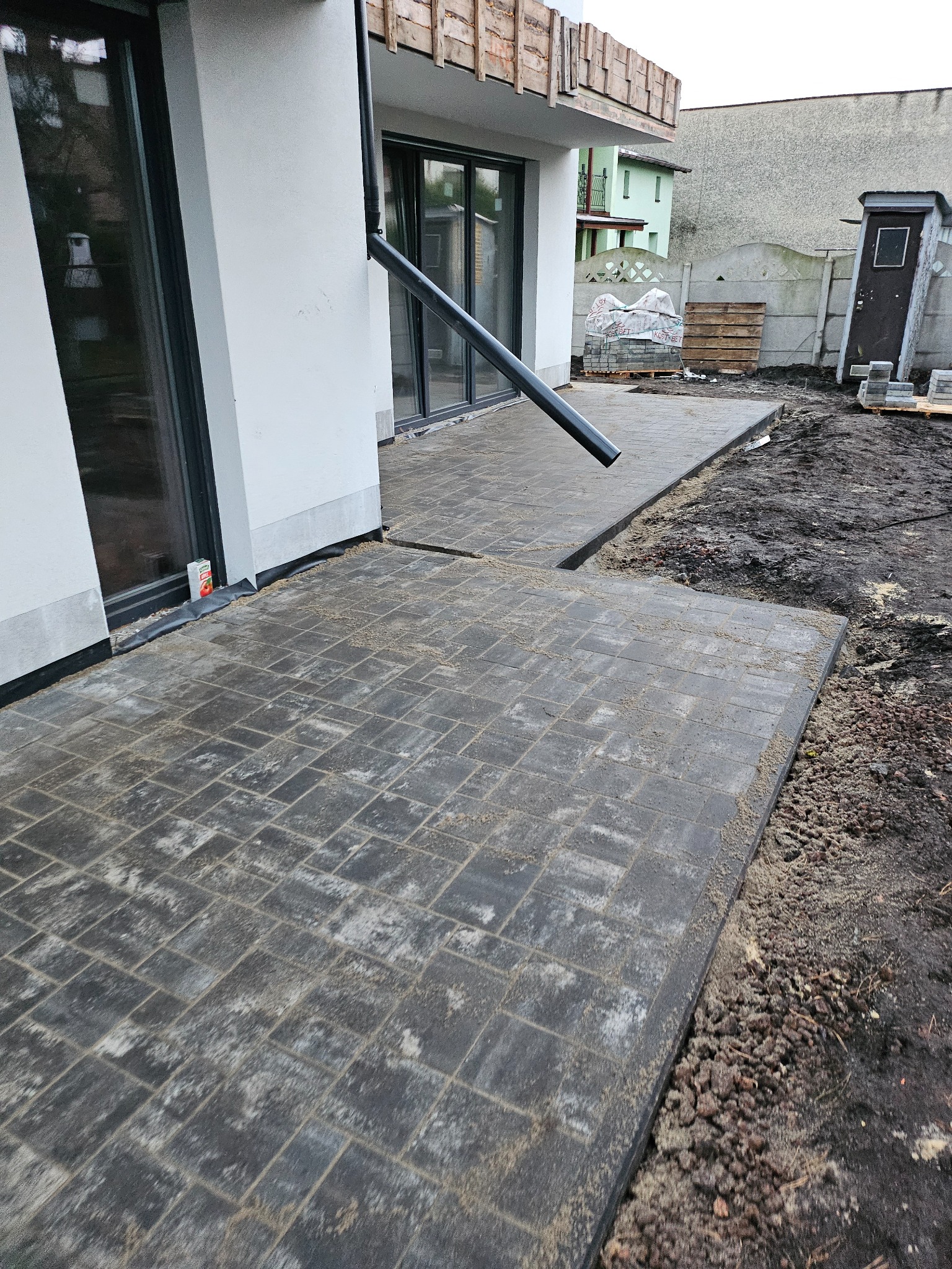 Ułożona, szara kostka brukowa przy białym budynku z rynną, widoczne palety z kostką w tle.