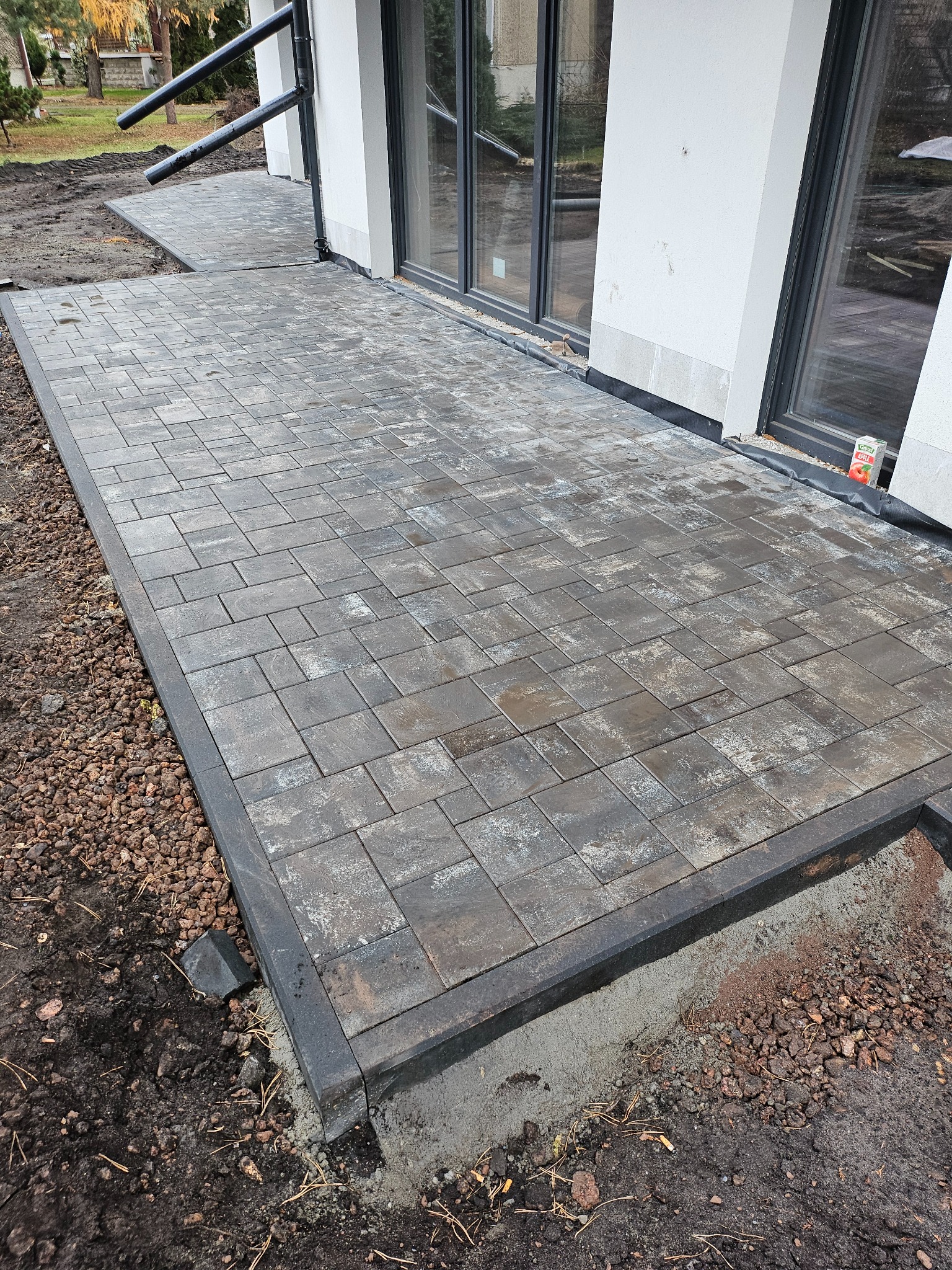 Ułożony chodnik z szarej kostki brukowej z ciemną obwódką przy białym budynku z dużymi oknami, widoczne elementy wykończeniowe i ziemia.