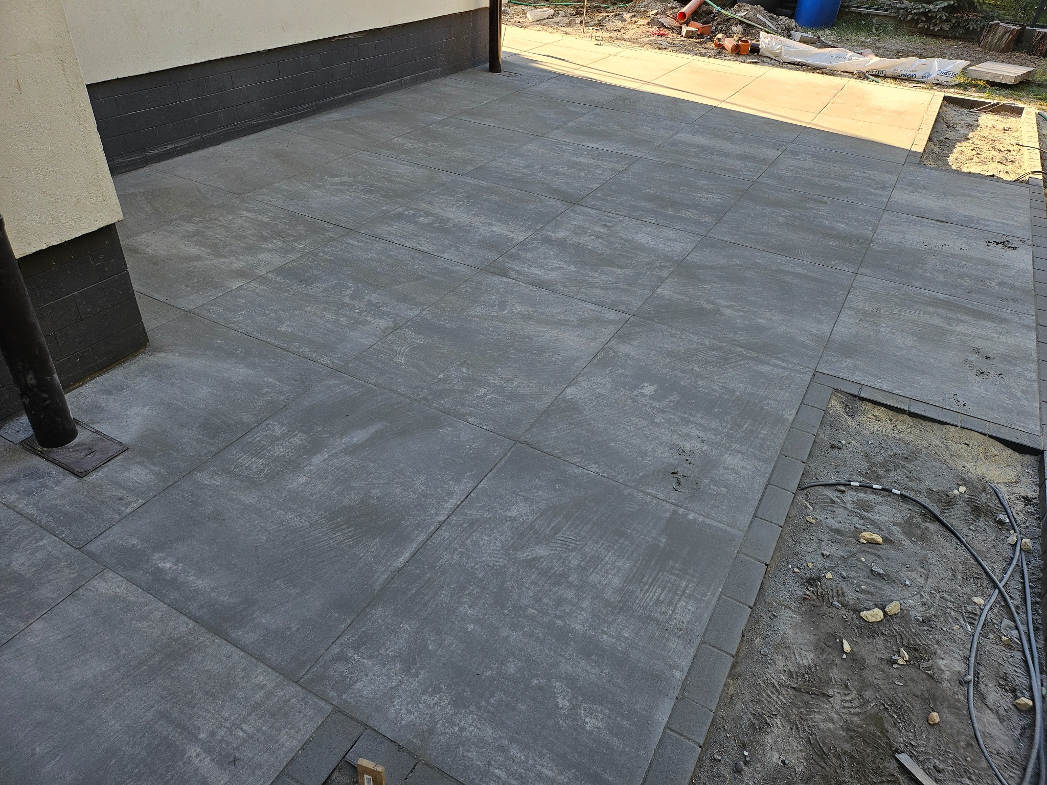 Świeżo ułożony, szary taras z kostki brukowej o dużej powierzchni, z widocznym fragmentem niedokończonej krawędzi i przygotowanym podłożem z kablami.