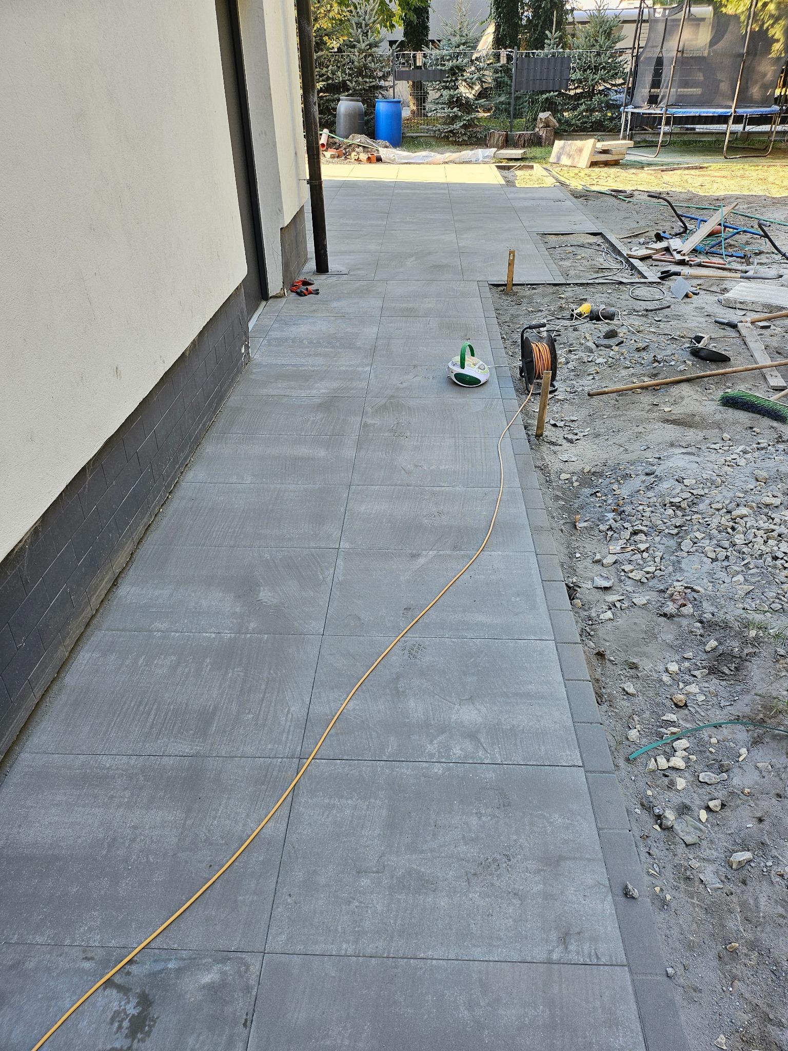Świeżo ułożona, szara kostka brukowa tworząca chodnik przy budynku, z widocznymi narzędziami i przewodem elektrycznym na powierzchni oraz niezakończonym fragmentem z piaskiem i kamieniami po prawej...