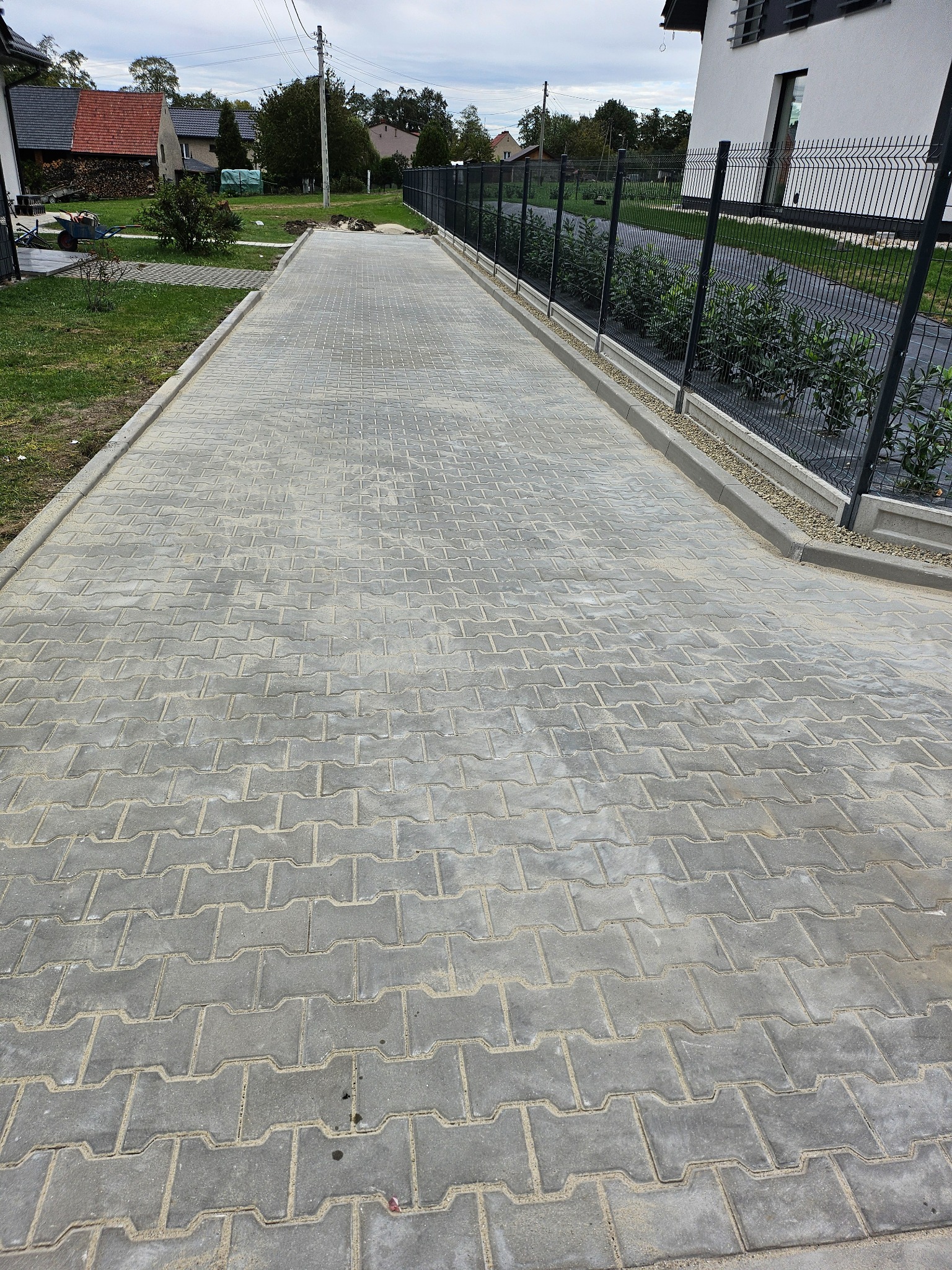 Długa, szara droga z kostki betonowej ułożona w zygzak, obrzeża z szarego betonu, czarne ogrodzenie panelowe po prawej stronie, trawnik po lewej, domy w tle.
