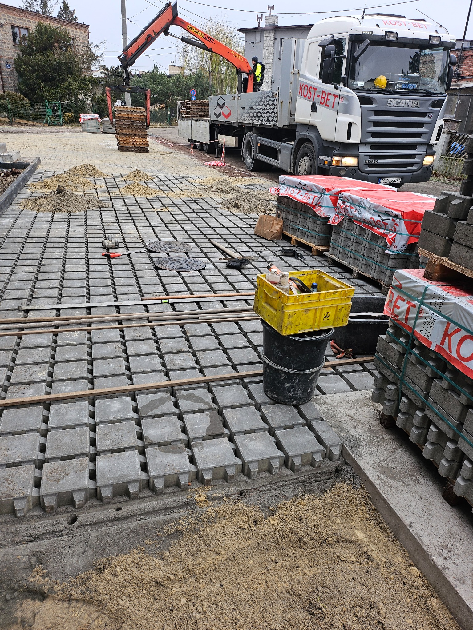 Układanie podbudowy pod nawierzchnię z prefabrykowanych elementów betonowych z użyciem dźwigu samochodowego; widoczne palety z kostką, narzędzia i piasek.