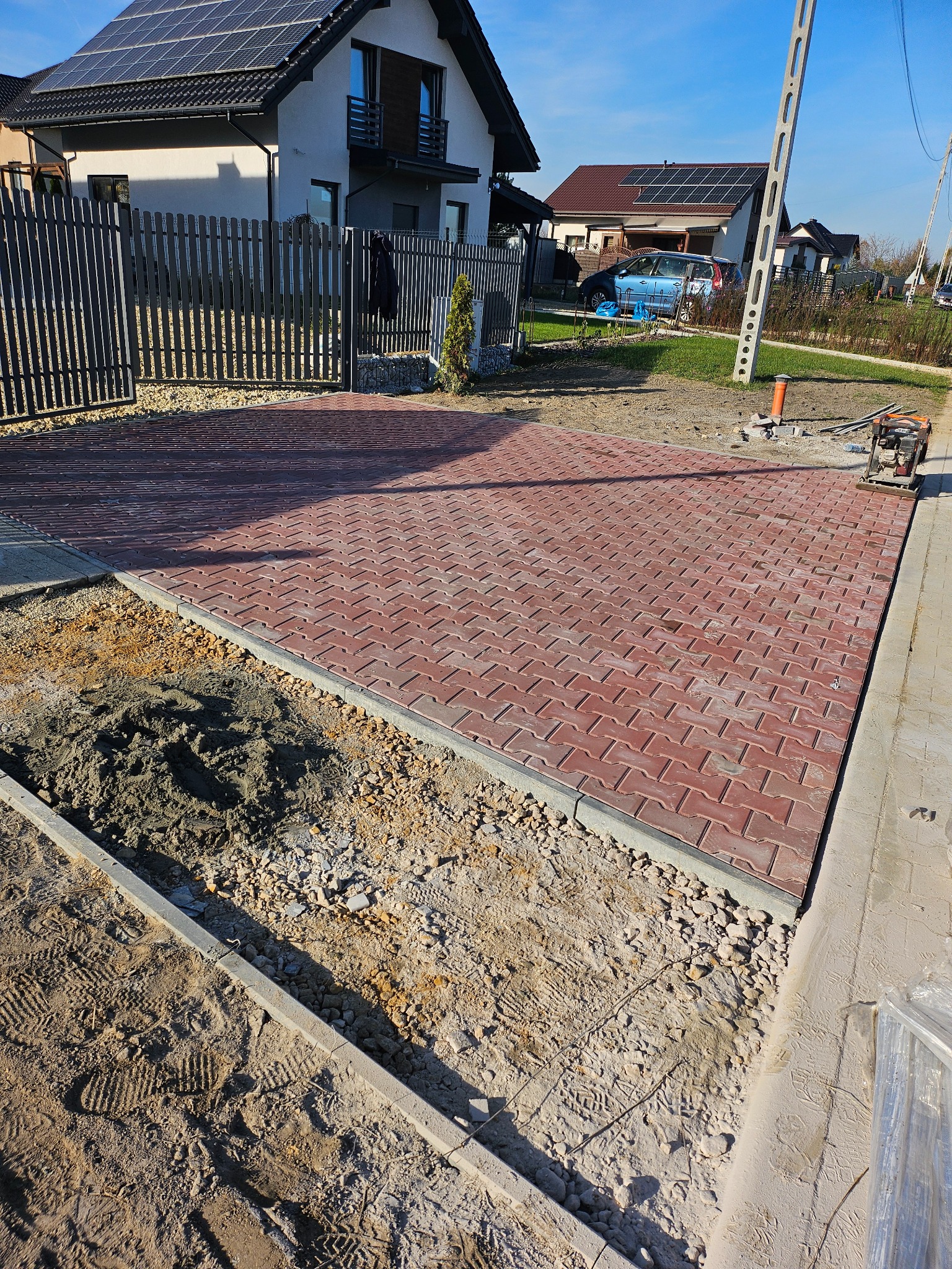 Częściowo ułożona, czerwona kostka brukowa na posesji domu jednorodzinnego, widoczna warstwa podsypki i ubijarka wibracyjna.