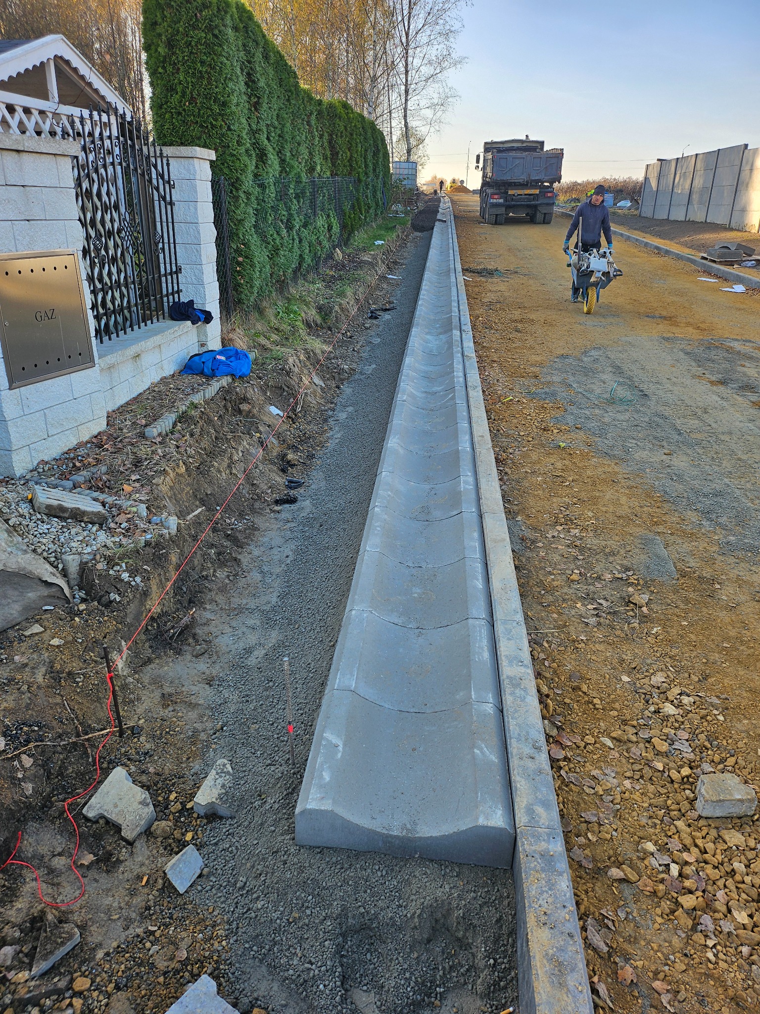 Montaż koryta odwadniającego z betonu, pracownik zagęszcza podłoże ubijarką wibracyjną, w tle ciężarówka i ogrodzenie posesji.