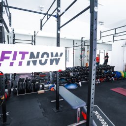 FitNow Kraków - studio treningów. Trener Personalny - Przestronna sala treningowa z dużą ilością sprzętu do ćwiczeń siłowych, w tle widoczni ludzie, w centralnej części baner z napisem FitNow.