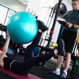 Kobieta ćwiczy z dużą, niebieską piłką fitness leżąc na macie, podczas gdy mężczyzna z clipboardem obserwuje i notuje postępy w siłowni z widocznymi hantlami i innym sprzętem.