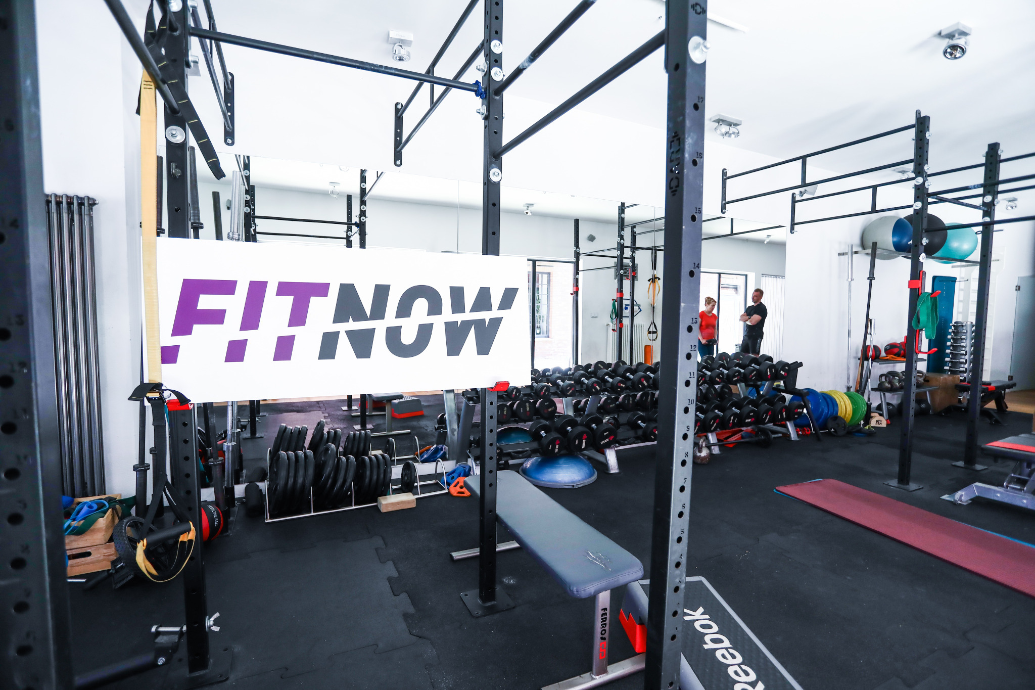 Przestronna sala treningowa z dużą ilością sprzętu do ćwiczeń siłowych, w tle widoczni ludzie, w centralnej części baner z napisem FitNow.