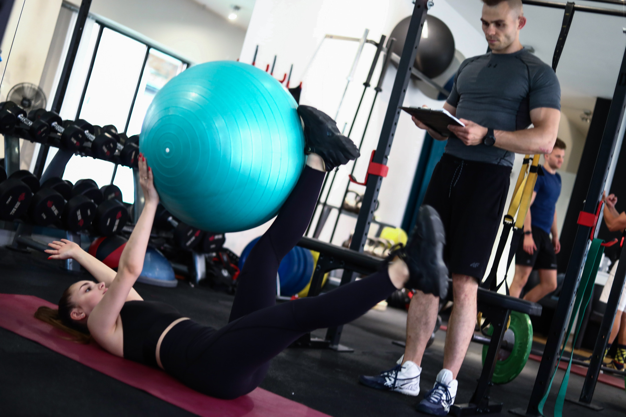 Kobieta ćwiczy z dużą, niebieską piłką fitness leżąc na macie, podczas gdy mężczyzna z clipboardem obserwuje i notuje postępy w siłowni z widocznymi hantlami i innym sprzętem.