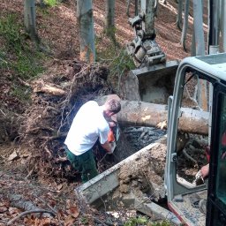 F.H.U Rafacz Szymon - Pracownik w białej koszulce tnie piłą spalinową korzeń drzewa w lesie, obok koparka z łyżką i kabina pojazdu z widocznym panelem sterowania.