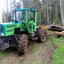 F.H.U Rafacz Szymon - Zielony traktor z przyczepą w błotnistym terenie leśnym, ciągnący ścięte pnie drzew