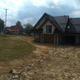 F.H.U Rafacz Szymon - Nowo budowany dom z czarnym dachem i szarymi kamiennymi elementami, widoczny teren po robotach ziemnych i stary budynek w tle.