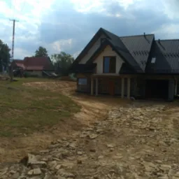 Nowo budowany dom z czarnym dachem i szarymi kamiennymi elementami, widoczny teren po robotach ziemnych i stary budynek w tle.