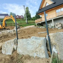 Żółta koparka przy budowie skalnego muru oporowego na nierównym terenie, z widokiem na drewniany dom z poddaszem i panele słoneczne w tle.