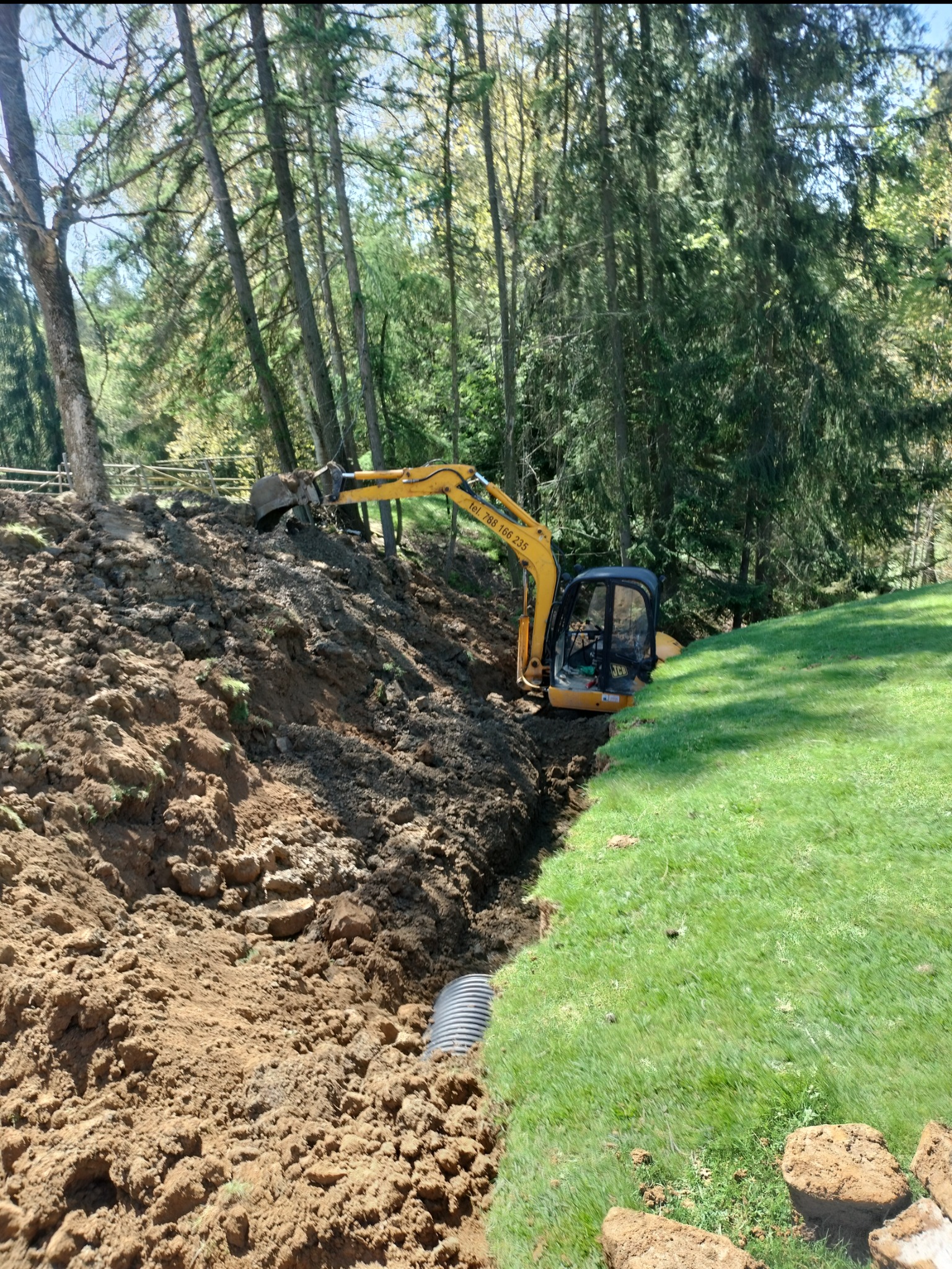 Żółta mini koparka w wykopie na skraju lasu, widoczna rura drenażowa w ziemi, świeżo wykopana ziemia i trawa.