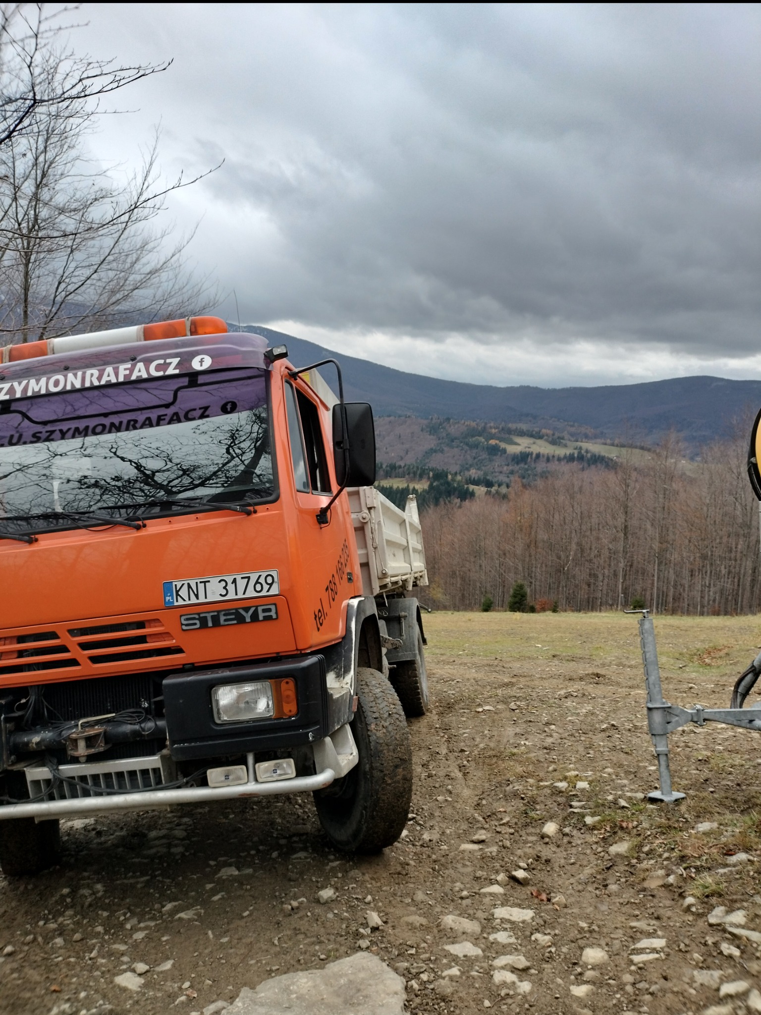 Pomarańczowa wywrotka Steyr z tablicą rejestracyjną KNT 31769, zaparkowana na nierównym, kamienistym terenie z widokiem na zalesione wzgórza pod pochmurnym niebem; na kabinie naklejka z nazwą firmy.