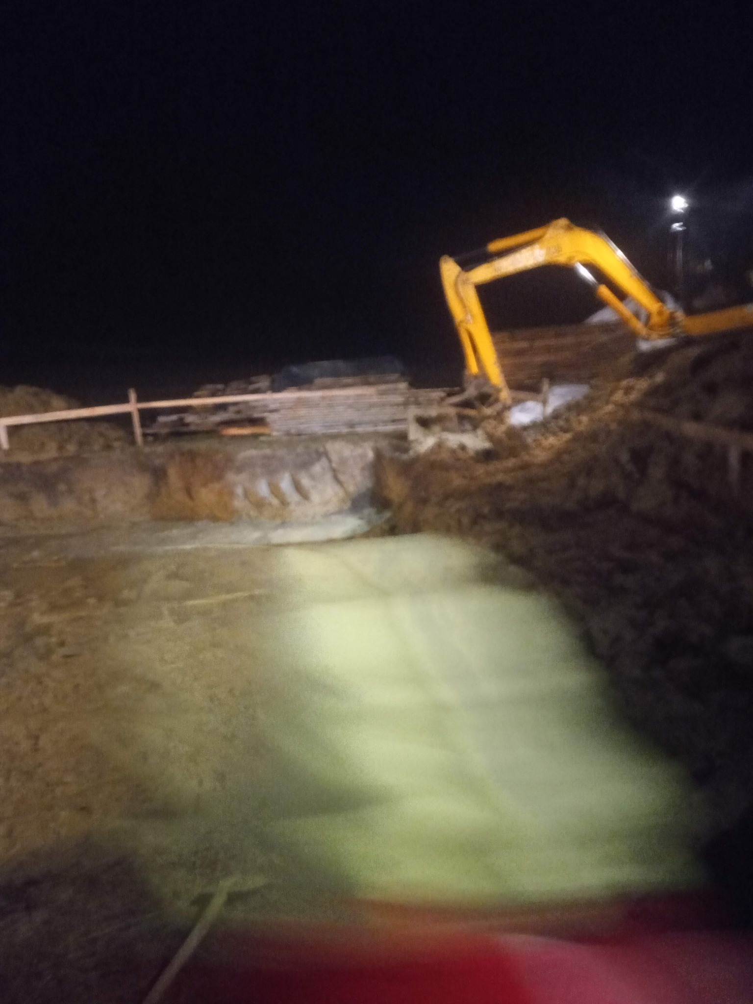 Żółta koparka na placu budowy nocą, oświetlona lampą, wykop pod fundamenty z widocznym szalunkiem.