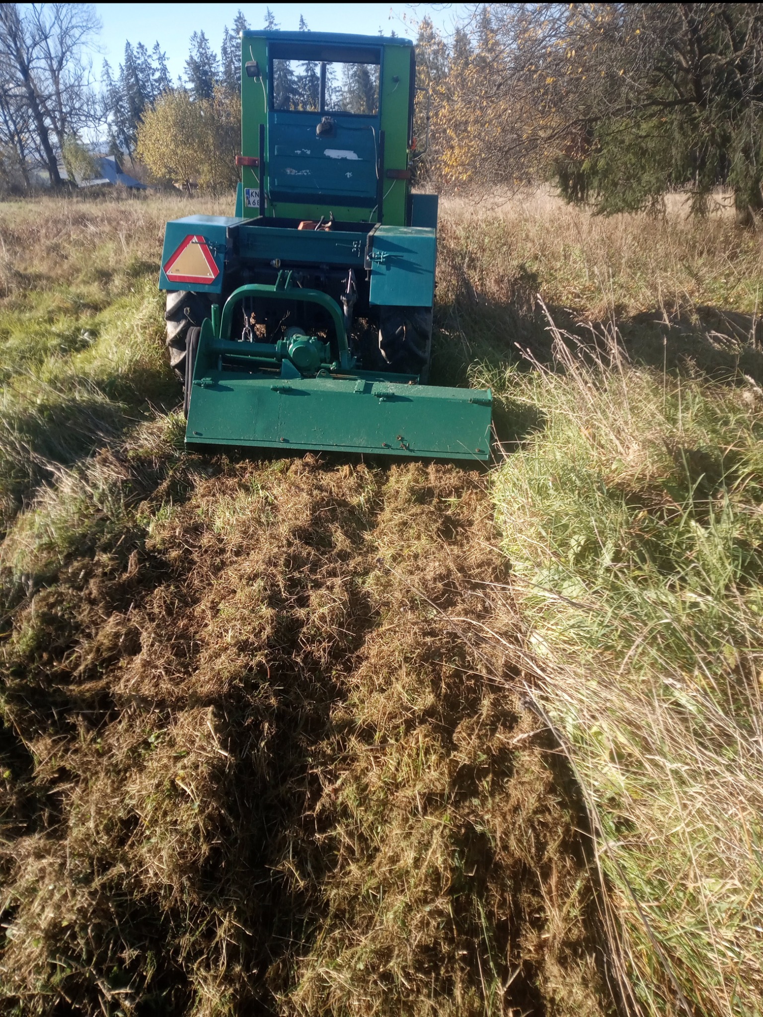 Zielony traktor z kosiarką rotacyjną tnący wysoką trawę na łące w słoneczny dzień, widok z tyłu.