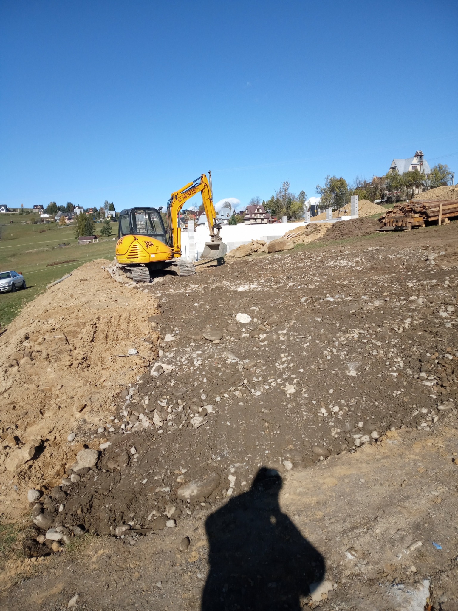 Żółta koparka JCB na nierównym, częściowo wyrównanym terenie budowy, z widocznymi fundamentami w tle i stosem drewna, w słoneczny dzień.