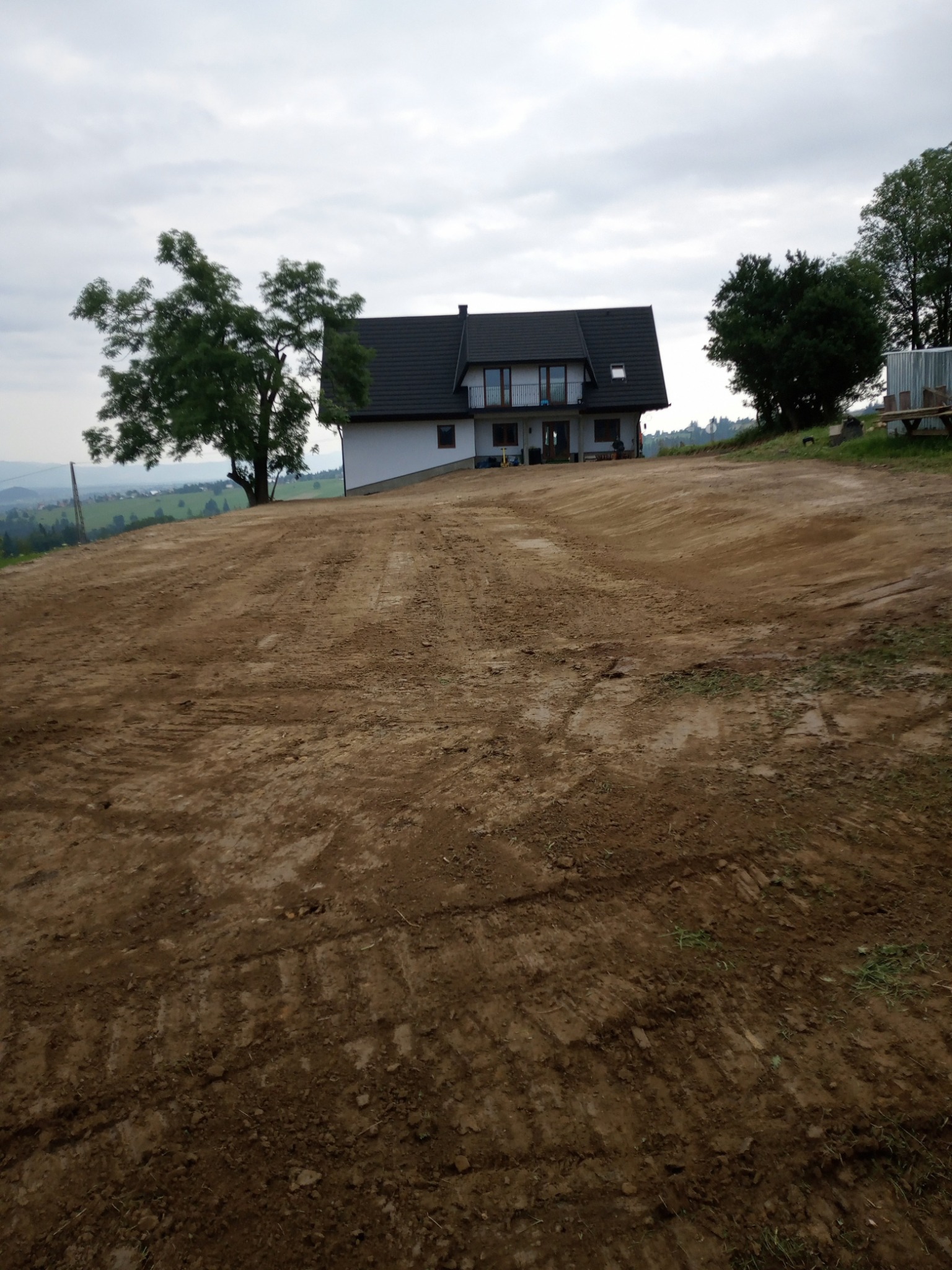 Przygotowany teren pod budowę domu, wyrównana powierzchnia ziemi z widocznymi śladami maszyn, w tle nowoczesny dom z balkonem i drzewa, pochmurne niebo.
